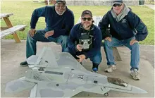 Three men kneeling behind a large model jet on a concrete surface.