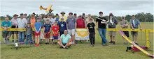 Group of people with model airplanes, smiling outdoors near a yellow fence.