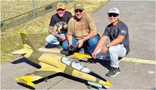 Three men kneeling on pavement with a model aircraft, smiling.
