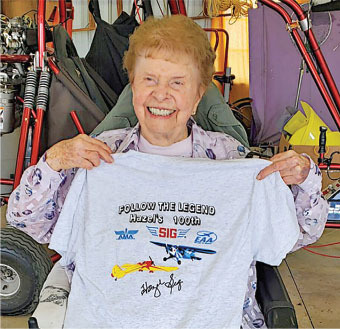 Elderly woman smiling, holding a shirt with planes and text "Follow the Legend, Hazel's 100th."