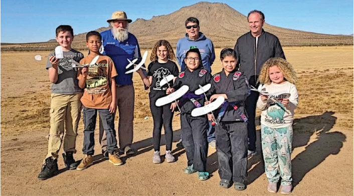 Group of people holding model planes in a desert landscape, mountain in background.
