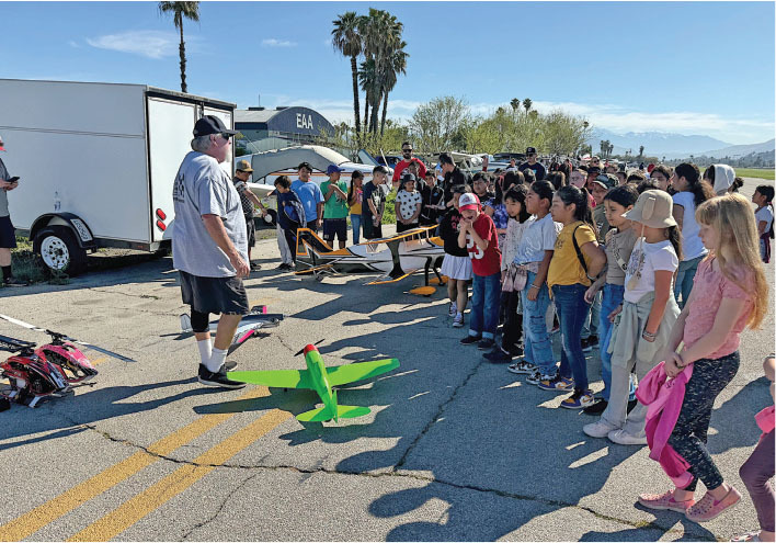 Man demonstrating model planes to a group of children on a sunny day.