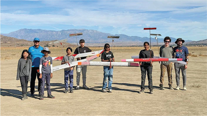 Group of people outdoors holding two long, colorful model planes. Mountains in the background.