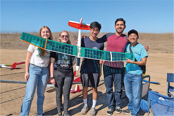 Five people smiling, holding a model plane outdoors under clear skies.