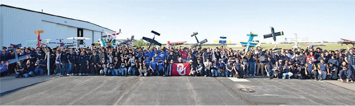 Large group with model planes and flags, outside an aircraft hangar on a sunny day.