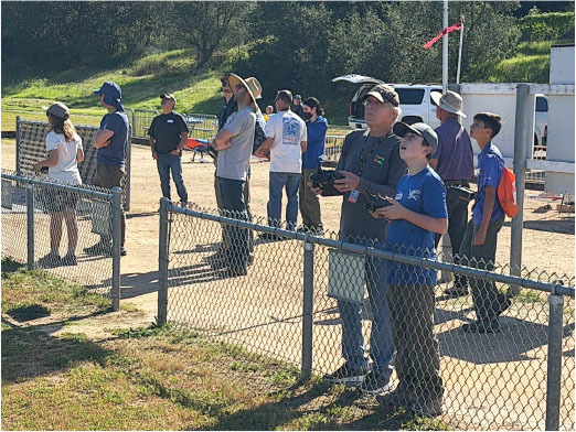 People watching and controlling drones by a fence under a sunny sky.
