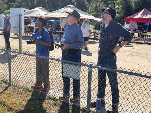 Three men standing outdoors, holding remote controls, behind a chain-link fence.