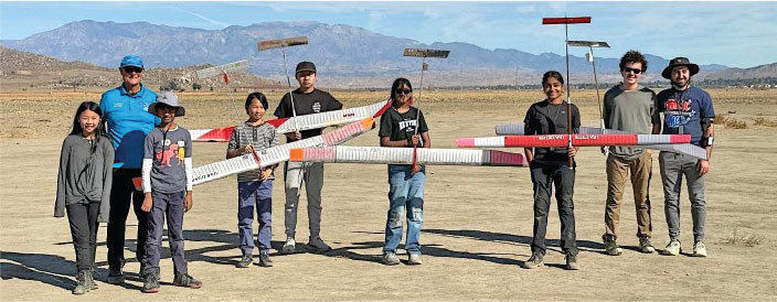 Eight people outdoors holding large model airplanes. Desert landscape, mountains in background.
