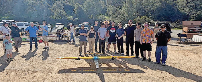 Group of people posing outdoors with a model plane on a sunny day.