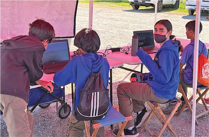 Children using laptops under a canopy, one holds a water bottle and wears a mask.