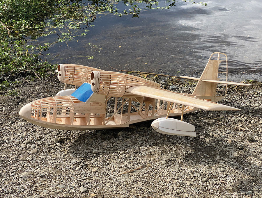 Wooden model seaplane on pebbled riverbank with blue cockpit accent.