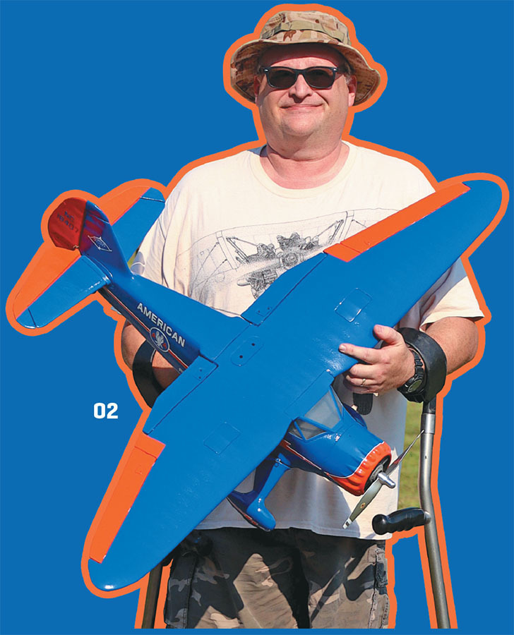 Man holding blue and orange model airplane, wearing hat and sunglasses.