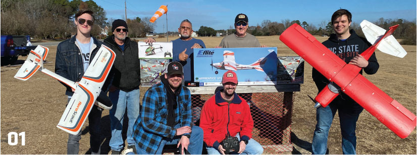 Group holding model planes, posing outdoors with a blue sky and grassy field.