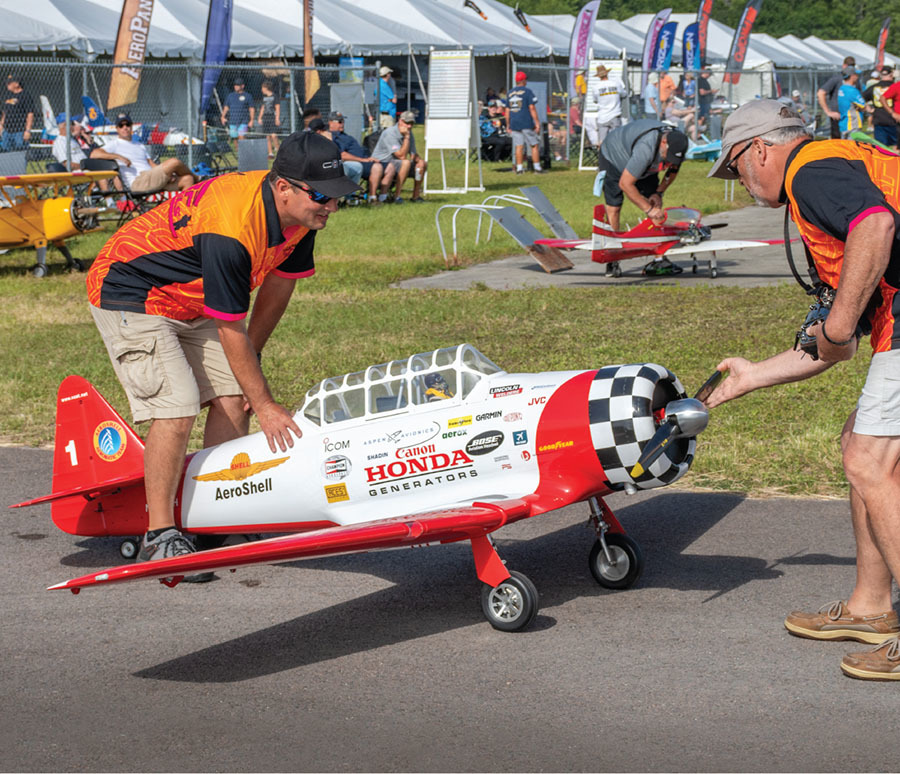 Two men in orange vests hold a scale model plane with sponsor logos.