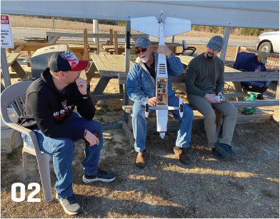 Three men seated, examining a model airplane outdoors.