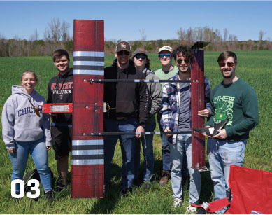 Group holding a model plane outdoors, number "03" at the bottom left.