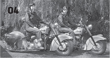 Two people in vintage gear on classic motorcycles under a tree.