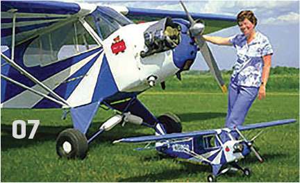 Woman with model plane next to real plane on grassy field, blue sky above.