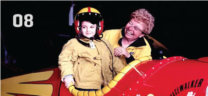 Child in a yellow jacket and helmet with an adult beside a red airplane nose, smiling.