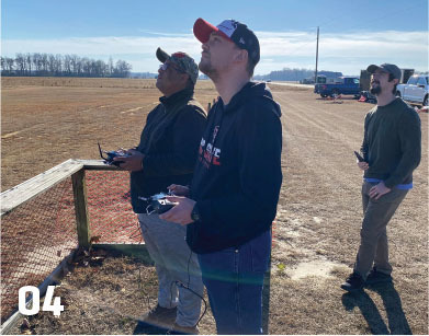 Three men flying drones in an open field, focused and holding controllers.