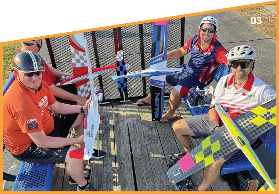 Four men with helmets holding model planes, seated on blue benches, outdoors.