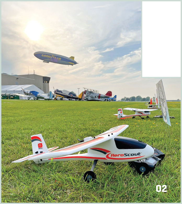 Model airplane on grass with Goodyear blimp in sky and vintage planes in background.