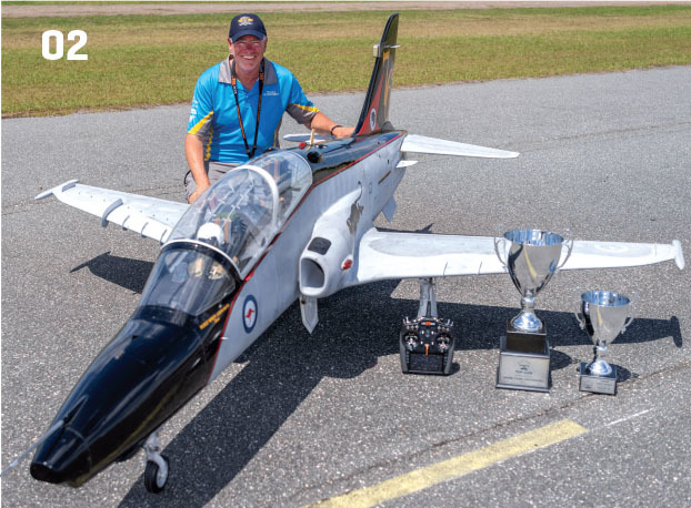 Man in blue shirt with model jet and trophies on asphalt.