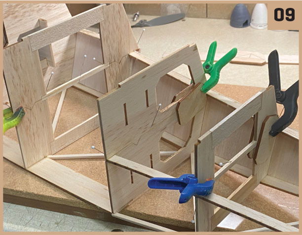 Wooden airplane model under construction, held with colored clamps.