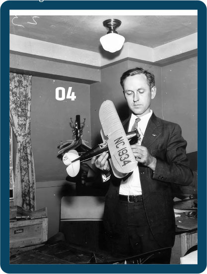 Man in suit examines model airplane labeled "NC 1934" in a room with patterned curtains.
