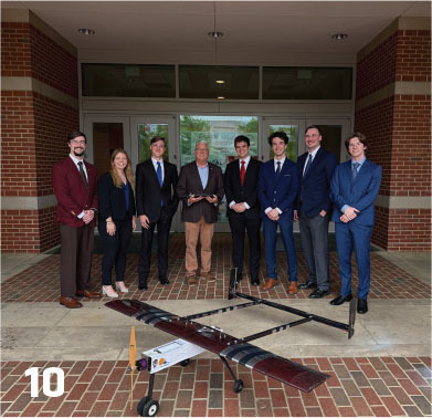 Eight people in formal attire stand behind a small model plane on a brick pathway.