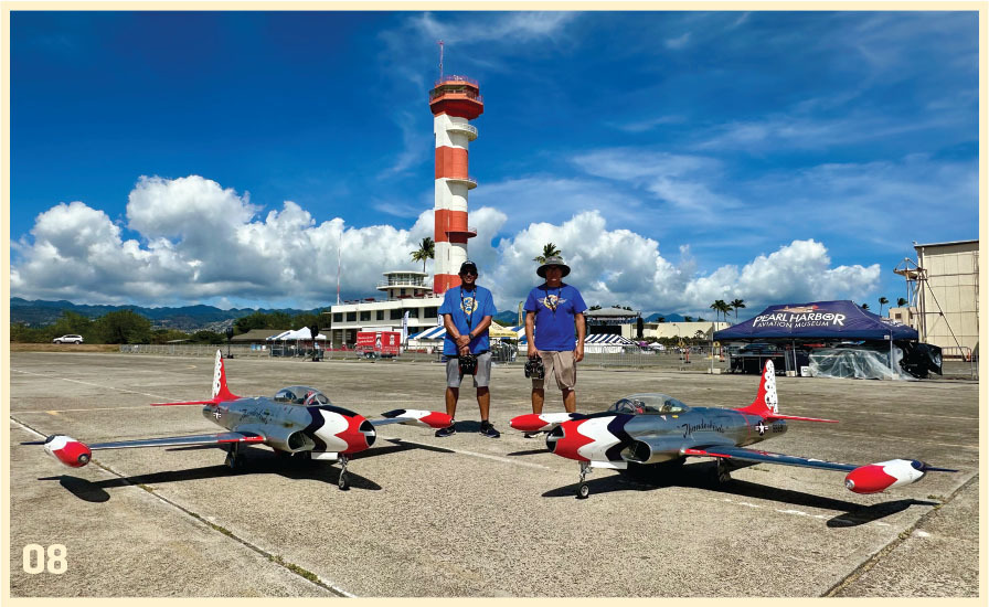 Two people with model jets at an airfield, red and white control tower behind.