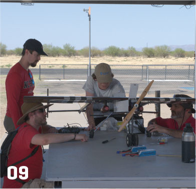 Four people with a drone on a table outdoors, number 09 in the corner.