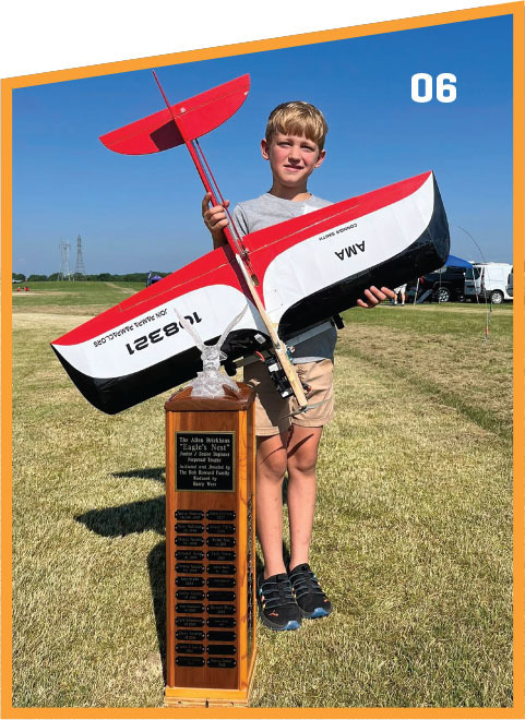 Boy holding model plane next to tall trophy on grassy field, blue sky background.