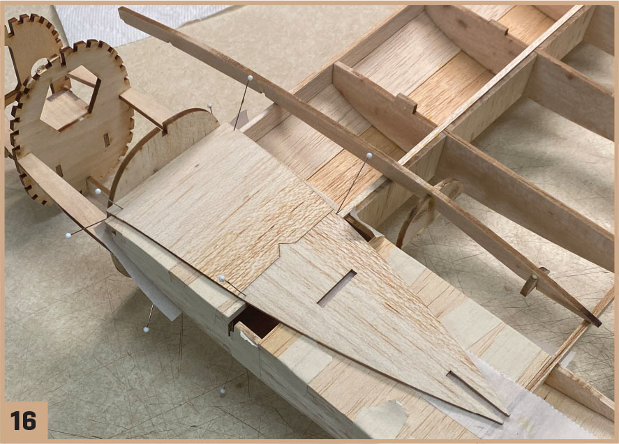 Wooden model airplane under construction with pins and gears; number 16 in the corner.