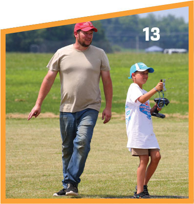 Man and boy flying a model aircraft in a grassy field, number 13 in corner.