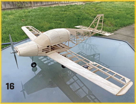 Wooden model airplane on a glass table, grass in background. Number 16 visible.