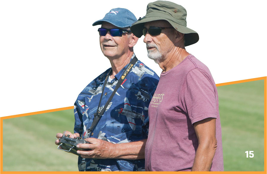 Two men outdoors, one holding a remote control, both wearing hats and sunglasses.