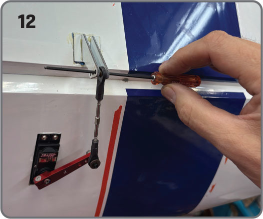 Hand using screwdriver to adjust remote control airplane part, number 12 shown.