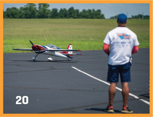 Man in cap watches model plane on tarmac, grassy field in background. Number 20 in corner.
