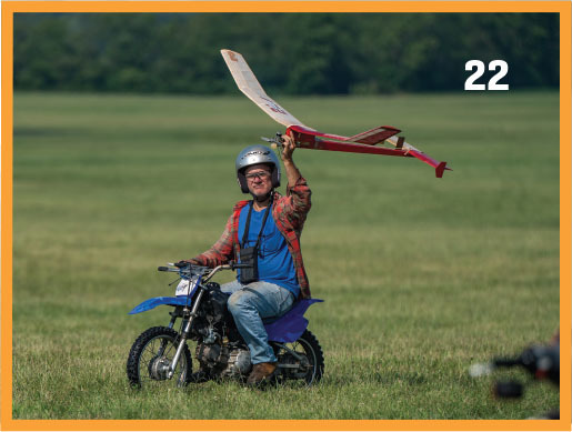 Man on motorbike holds model plane in grassy field, number 22 in corner.