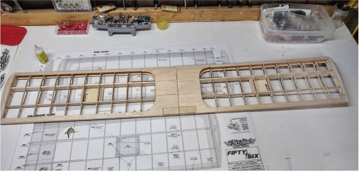 Wooden model airplane wing frame on a workbench, with plans labeled "Fifty Six".