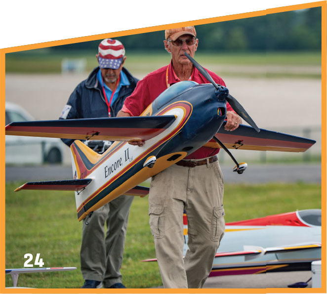 Two men carrying a model plane labeled "Encore II" on a grassy field.