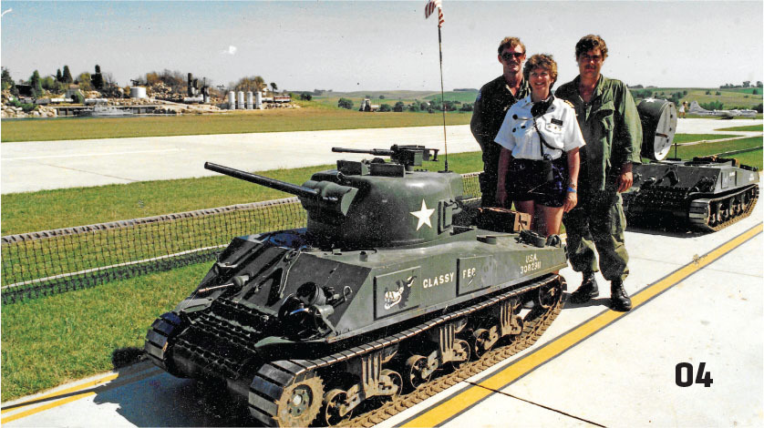 Three people stand by a small military tank on display outdoors.