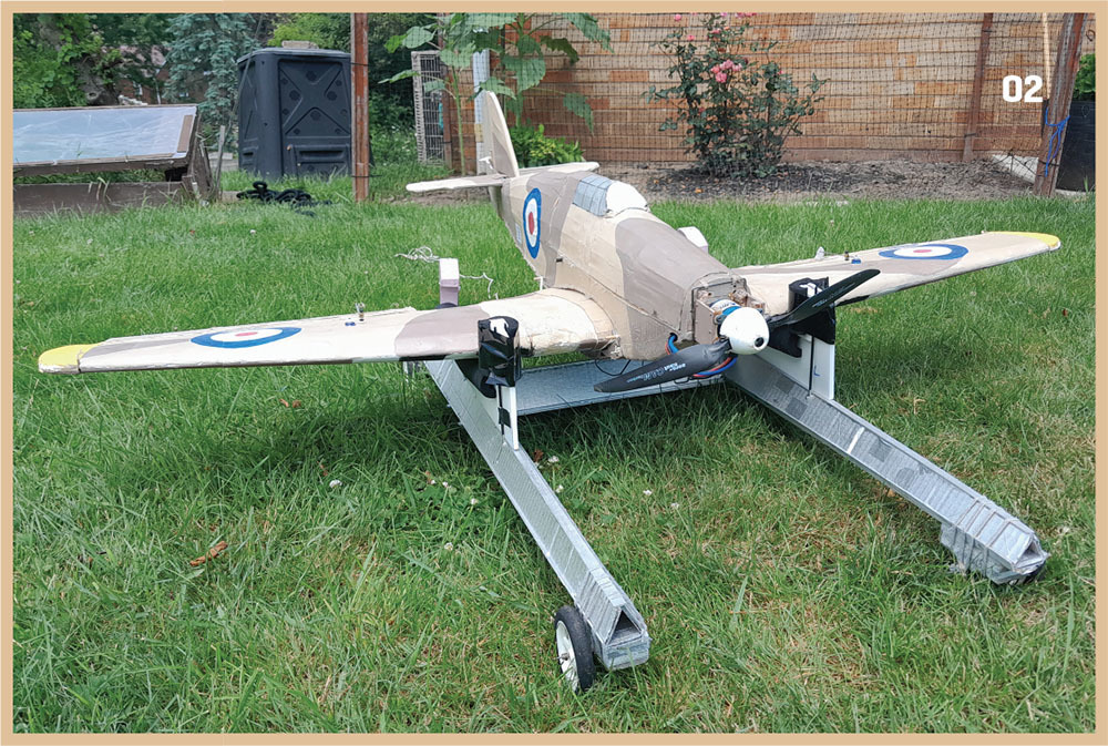 Toy model plane on grass with RAF roundels on wings, surrounded by garden scenery.