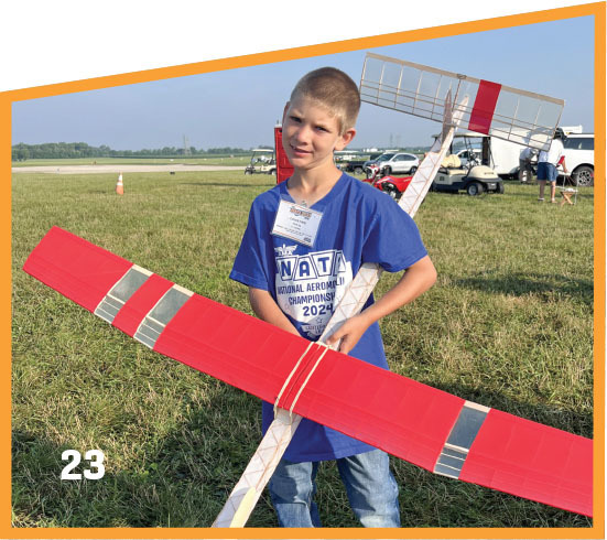 Young boy in a blue T-shirt holds a large red model plane outdoors, text reads "NATs 2024".