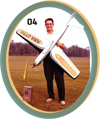 Man holding model airplane with "AMA-2711" text, standing on grassy field.