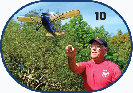 Man in red shirt launching a model airplane outdoors, number 10 in background.