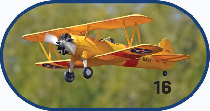 Yellow biplane with U.S. Navy markings, number 16, mid-flight against a blurred green background.