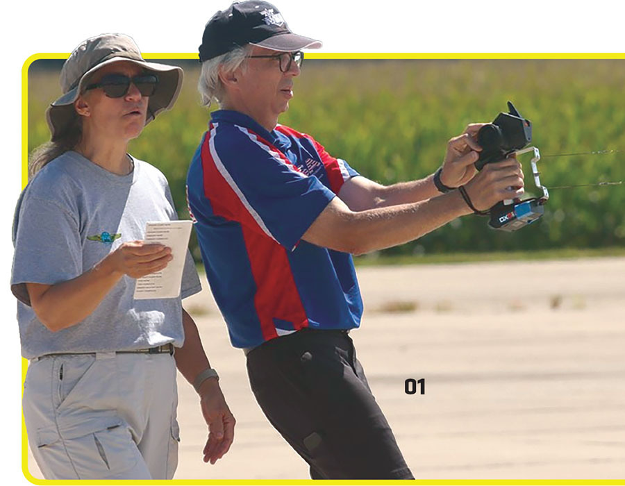 Man flying model plane with controller; woman beside him holding a paper.