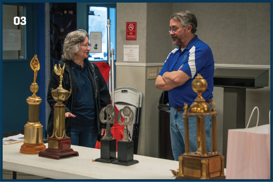 Two people talking near a table with several large trophies.